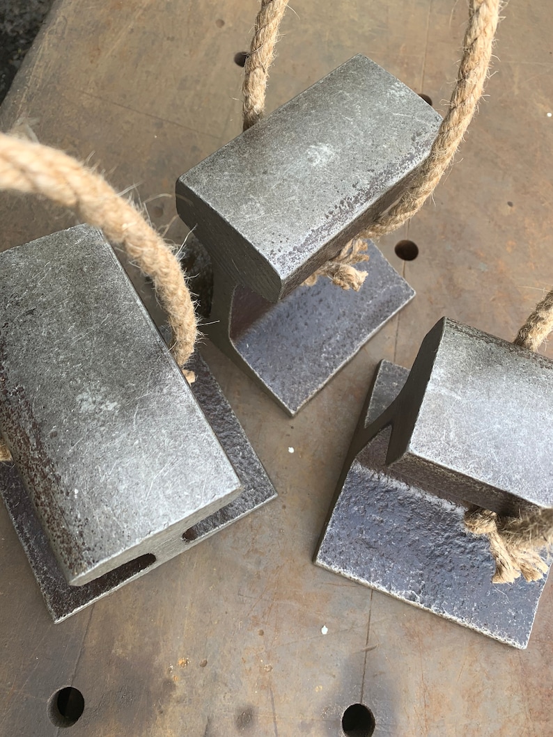 May include: Four weathered metal doorstops with attached rope handles rest on a metal surface. The doorstops are rectangular with a T-shaped base. The rope is a natural tan color.