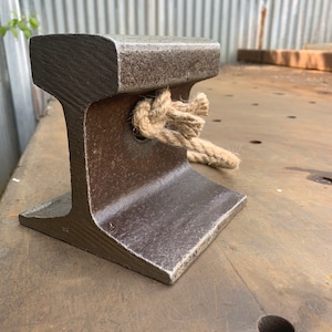 May include: A weathered, dark gray metal railroad tie section with a rope tied through a hole. The tie has an I-beam shape and sits on a brown surface. The rope is a natural beige color.