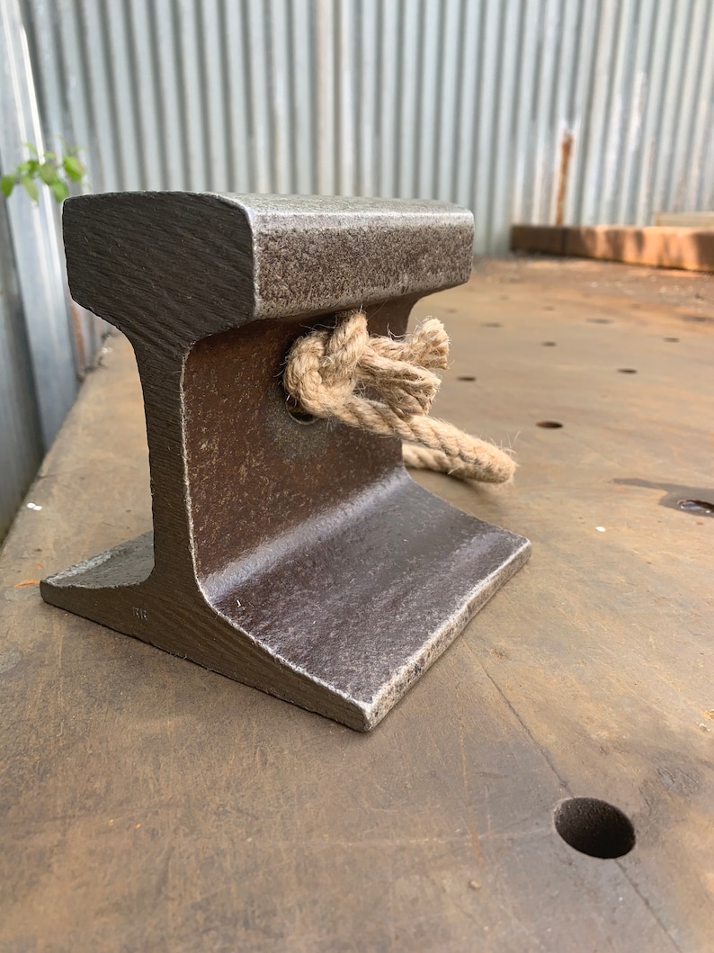 May include: A weathered, dark gray metal railroad tie section with a rope tied through a hole. The tie has an I-beam shape and sits on a brown surface. The rope is a natural beige color.