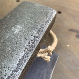 May include: Close-up of a weathered, rectangular metal object with a rope handle. The metal has a textured, aged appearance, with visible imperfections. The rope is a natural beige color, adding a rustic touch to the design. The object appears to be a decorative or functional item.