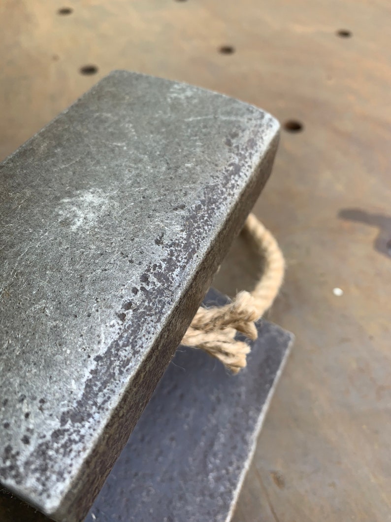 May include: Close-up of a weathered, rectangular metal object with a rope handle. The metal has a textured, aged appearance, with visible imperfections. The rope is a natural beige color, adding a rustic touch to the design. The object appears to be a decorative or functional item.