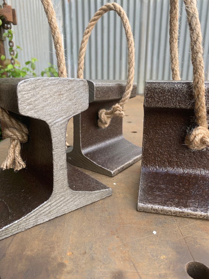May include: Three metal bookends with a rustic, industrial design. Each bookend is shaped like a section of a railroad track and features a natural rope handle. The bookends have a weathered, textured appearance with shades of gray and brown.