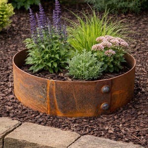 May include: A circular, weathered metal planter filled with various plants. The plants include purple flowering spikes, green grass, and pink blooms. The planter is surrounded by brown mulch and bordered by stone pavers, creating a rustic garden bed.