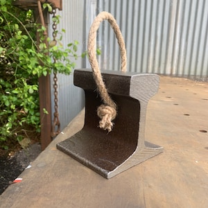 May include: A dark gray, industrial-style doorstop made from a section of steel rail. It features a natural rope handle tied in a knot. The doorstop is designed to stand upright and has a sturdy, rectangular base.