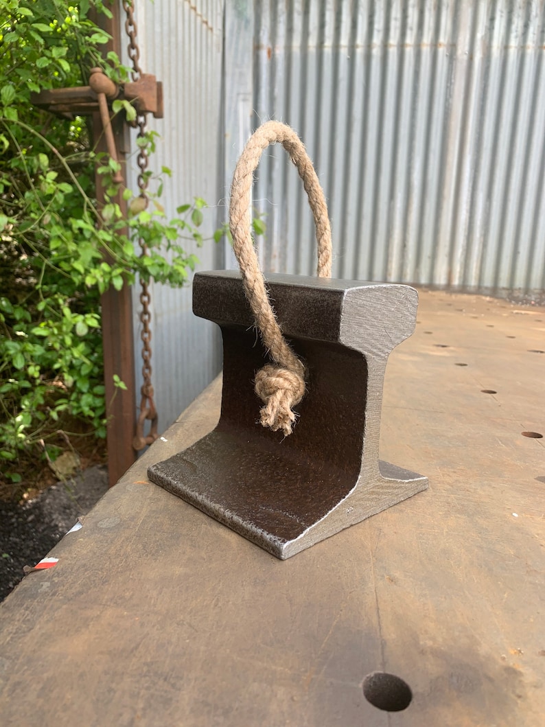 May include: A dark gray, industrial-style doorstop made from a section of steel rail. It features a natural rope handle tied in a knot. The doorstop is designed to stand upright and has a sturdy, rectangular base.