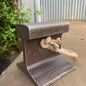 May include: A weathered, dark brown metal railroad tie section with a rope tied through it. The tie has a textured surface and a flat top. The rope is a natural beige color and is knotted in the center.