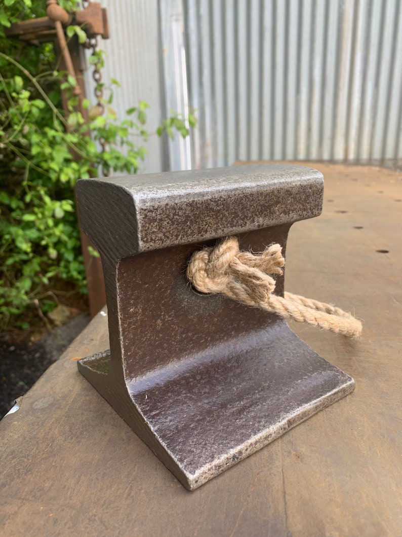 May include: A weathered, dark brown metal railroad tie section with a rope tied through it. The tie has a textured surface and a flat top. The rope is a natural beige color and is knotted in the center.