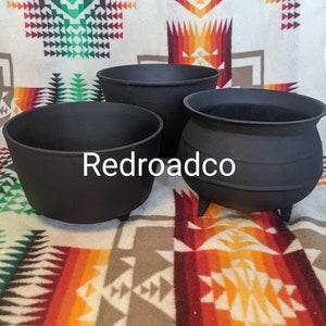 May include: Three black cast iron pots with three legs each. The pots are on a colorful blanket with a geometric pattern.