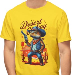Puede incluir: Una camiseta amarilla con una imagen de dibujos animados de un lagarto con un sombrero de vaquero y sosteniendo una pistola. El texto "Desert Cowboy" está impreso en la camiseta.