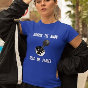 Op de afbeelding: Een koninklijk blauw t-shirt met een witte afbeelding van een autoversnellingspook en de tekst "Workin' the Knob Gets Me Places".