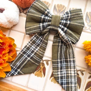 May include: An olive green plaid bow with long tails. The bow features a classic black and white check pattern. The bow is made of a soft fabric and is displayed on a decorative tile surface, with orange and white pumpkins and yellow flowers.