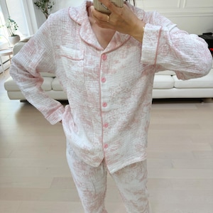 Puede incluir: Un conjunto de pijama de manga larga con estampado rosa y blanco y una parte superior con botones. La parte superior tiene cuello y un bolsillo en el pecho.