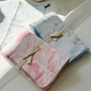 May include: Two sets of folded fabric baby washcloths. One set is pink with a white pattern and the other is blue with a white pattern. Both sets are tied with a white ribbon.