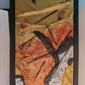 May include: Abstract art piece with a gold, silver, and red color scheme. The artwork is made of various materials, including fabric, foil, and sticks. The sticks are arranged in a criss-cross pattern.
