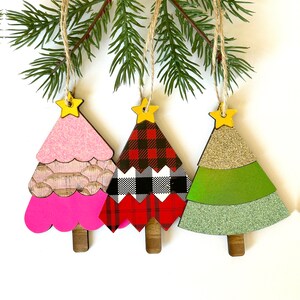 May include: Three wooden Christmas tree ornaments with different patterns. The first tree is pink with a white and pink pattern, the second tree is red and black plaid, and the third tree is green with a gold glitter pattern.