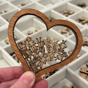 Puede incluir: Decoración de madera en forma de corazón con un centro transparente, con una familia y las palabras "Happy Mother's Day". El corazón se sostiene en una mano, con otras formas de madera visibles en el fondo.