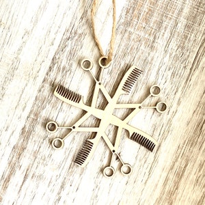 May include: A wooden snowflake ornament with a design of hairdressing tools, including combs and scissors. The ornament is hanging from a string.