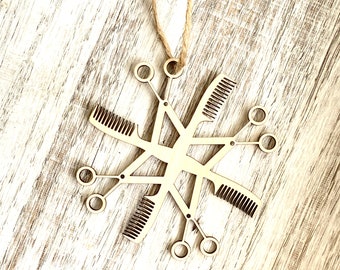 Op de afbeelding: Een houten sneeuwvlok ornament met een ontwerp van kappersgereedschap, waaronder kammen en scharen. Het ornament hangt aan een touwtje.