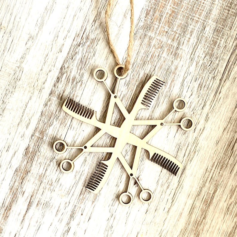 May include: A wooden snowflake ornament with a design of hairdressing tools, including combs and scissors. The ornament is hanging from a string.