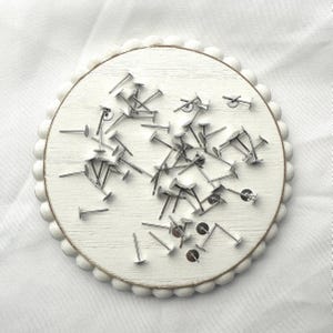 May include: A collection of silver-colored earring posts and backs scattered on a white, round, wooden surface with a scalloped edge. The posts have a flat, circular top and a straight post.