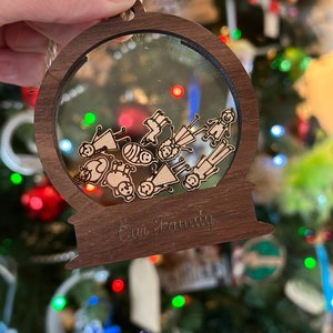 Può includere: Un ornamento natalizio in legno a forma di palla di neve con una cupola di plastica trasparente. La cupola contiene un disegno di una famiglia tagliato al laser con le parole "Our Family" incise sulla base.