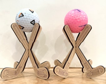 Puede incluir: Dos soportes de palo de golf de madera con una bola de golf blanca en uno y una bola de golf rosa en el otro.