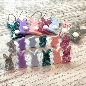 May include: A set of 14 bunny-shaped earrings in various colors, including pink, purple, blue, green, and white. Some earrings are glitter-covered, while others have a solid color. The earrings are perfect for adding a touch of whimsy to any outfit.