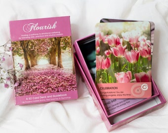 May include: A pink box with a deck of cards titled "Flourish: Finding a pathway to emotional wellbeing by following wise, practical and intuitive wisdom." The box contains a 52 card deck and guidebook. The cards feature a pink flower theme.