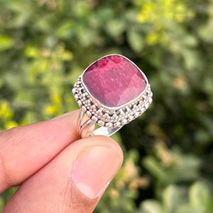 Op de afbeelding: Een zilveren ring met een vierkante, dieproze edelsteen gezet in een geperlde zetting. De ring zit aan een vinger.