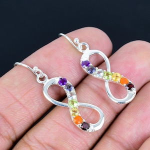 Op de afbeelding: Een paar zilverkleurige oorbellen met een oneindigheidssymbool. Elke oorbel heeft een reeks kleurrijke edelstenen, waaronder paars, blauw, groen, geel, oranje en rood, langs de curve van het oneindigheidssymbool.