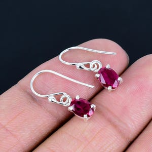 Puede incluir: Un par de pendientes colgantes de plata con piedras preciosas rojas oscuras de forma ovalada. Las piedras están engastadas en un bisel de plata y suspendidas de un gancho simple.