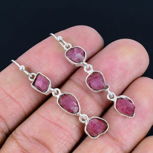 May include: Dangle earrings with raw, pink gemstones set in silver. Each earring has three stones of different shapes, linked by silver. The earrings are shown against a dark background. The gemstones are a vibrant pink.