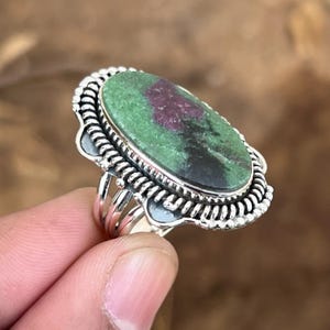 Peut inclure: Une bague en argent avec une pierre ovale verte, rose et noire sertie dans une bélière en argent détaillée. La bague a une bande torsadée.