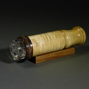 May include: A cylindrical kaleidoscope featuring a light-colored, figured wood body with a dark wood ring near the front. The front end has a clear glass lens, and the body displays a small, dark, handwritten signature. It rests on a rectangular dark wood base.