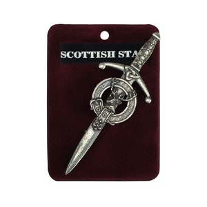 May include: A silver-colored Scottish dirk pin with a decorative hilt and blade, featuring a circular emblem with a stag's head. The pin is displayed on a burgundy velvet card with the text "SCOTTISH STA".
