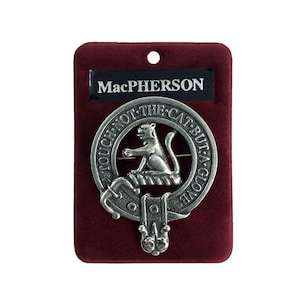 May include: A silver-coloured MacPherson clan crest pin featuring a cat on a rope design. The pin is displayed on a red velvet background. The text on the pin reads "Touch not the cat but a glove".