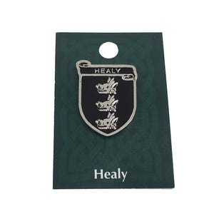 May include: A silver-colored Healy family crest pin with a black background, featuring three stylized animal heads. The word "HEALY" is displayed at the top of the crest, and the name "Healy" is printed at the bottom. The pin is mounted on a green card.