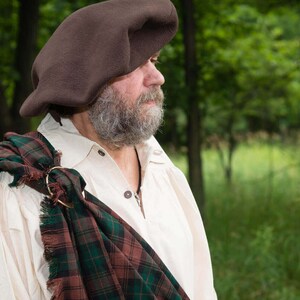 May include: A man wearing a brown beret and a white shirt with a green, brown, and red plaid kilt draped over his shoulder.