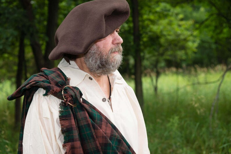 May include: A man wearing a brown beret and a white shirt with a green, brown, and red plaid kilt draped over his shoulder.