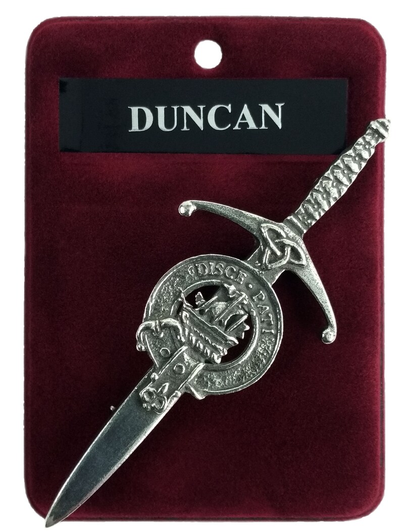 May include: Silver-toned kilt pin featuring a sword design with a Celtic knot and a circular crest with the text "DISC. ET. FATI" and a ship. The pin is displayed on a red velvet background with a black label that reads "Duncan".