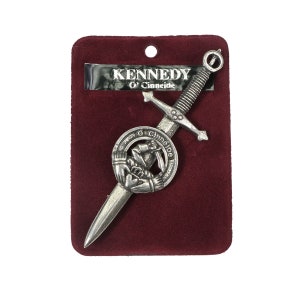 May include: A silver-colored Kennedy clan crest kilt pin, featuring a sword design with a circular emblem. The pin is displayed on a burgundy velvet backing, with the name "KENNEDY O' Cinneide" printed on a label.