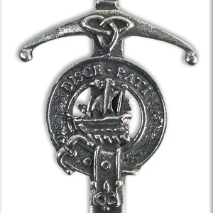 May include: A silver-toned kilt pin in the shape of a sword with a Celtic knot design. The sword is attached to a round shield with the words "DISCE. PATRI" and a ship design.