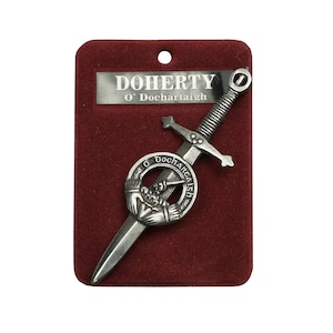 May include: A silver-colored kilt pin with a sword design and a circular crest featuring a hand holding a heart. The pin is displayed on a red velvet background with the text "DOHERTY O'Dоchаrtaigh" on a silver plaque.