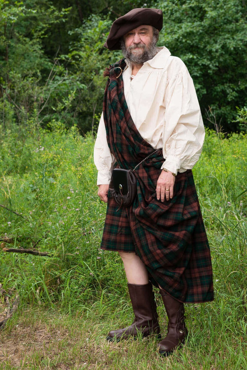 May include: A man wearing a brown hat and a white shirt with a plaid kilt. He is standing in a grassy field.