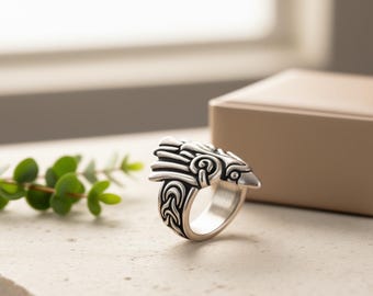 May include: A silver ring with a bird-like design. The ring showcases a detailed bird's head with intricate patterns and a polished band. The ring is presented on a light surface with a blurred background.