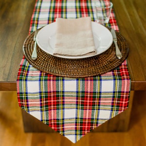 May include: A white and red plaid table runner with a brown woven charger, a white plate, and a beige napkin. The table runner is on a wooden table.