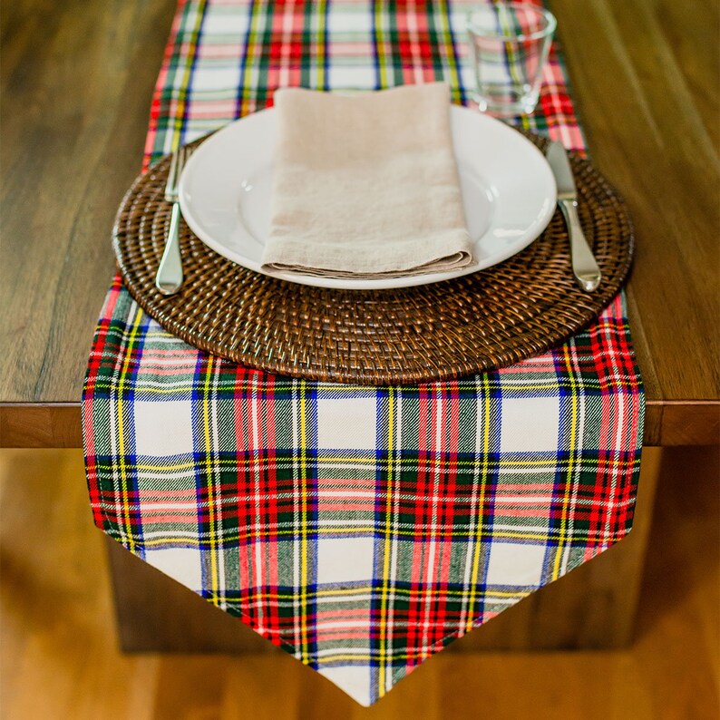 May include: A white and red plaid table runner with a brown woven charger, a white plate, and a beige napkin. The table runner is on a wooden table.