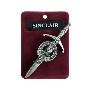 May include: A silver kilt pin shaped like a sword, featuring a circular emblem and the inscription "COMMIT THY WORK TO GOD." The pin is presented on a burgundy velvet card, with the name "SINCLAIR" printed on a black label.