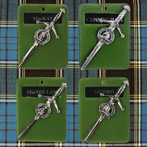 May include: Four silver-colored Scottish clan kilt pins with green velvet backing. The pins feature a sword with a circular crest and the clan name: MacKay, Cunningham, MacMillan, and Munro.