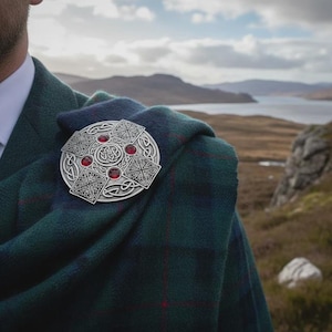 May include: A silver-toned Celtic brooch with red accents, pinned to a green and blue plaid shawl. The brooch features a circular design with intricate knotwork and square patterns. The background shows a scenic landscape with mountains and a body of water.
